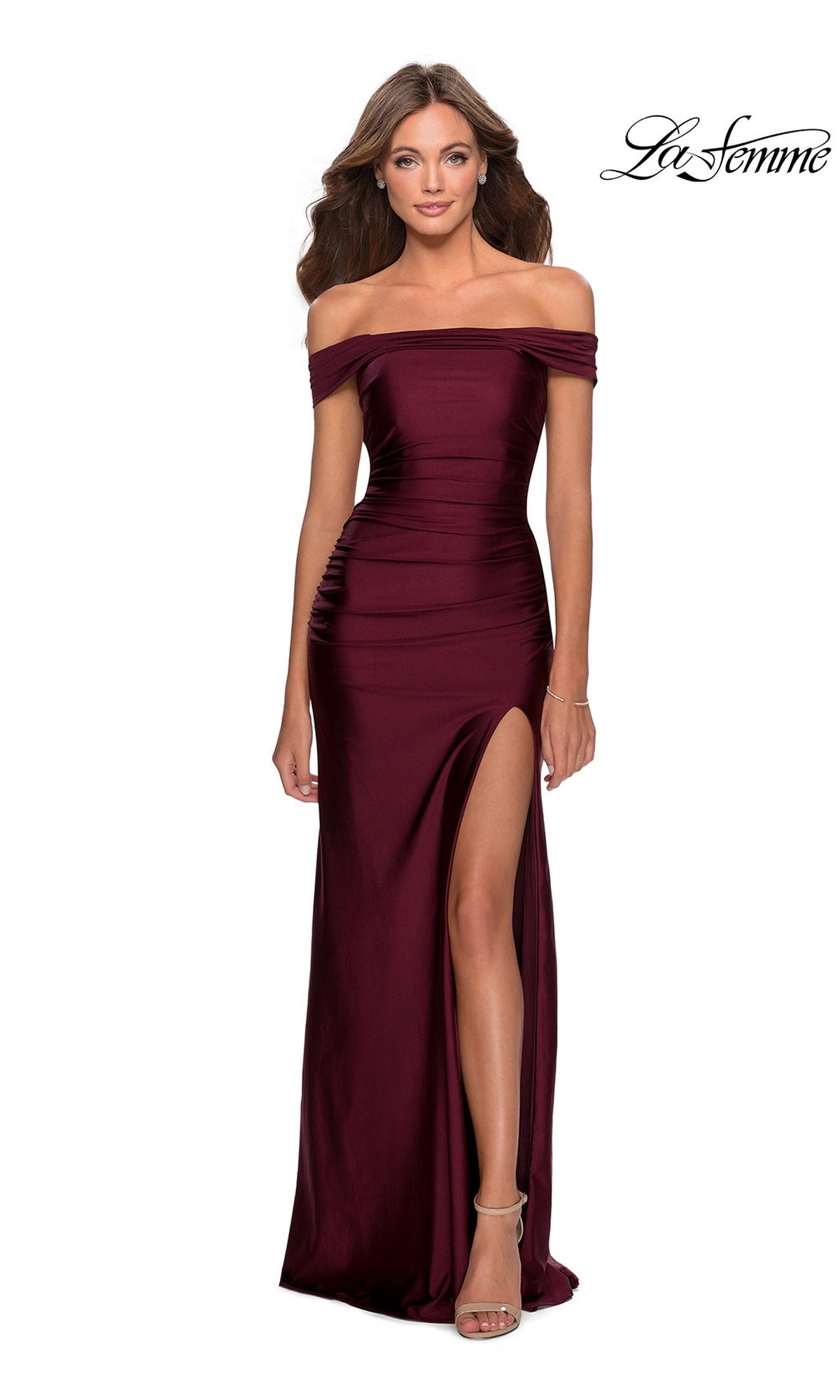 A woman wears the La Femme Strappy-Back Tight Long Prom Dress 28506 in burgundy, featuring an off-the-shoulder neckline, thigh-high slit, and strappy heels. The